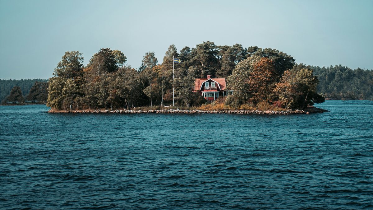 Bild på en liten ö med hus och flaggstång med svenska flaggan, i skärgården.