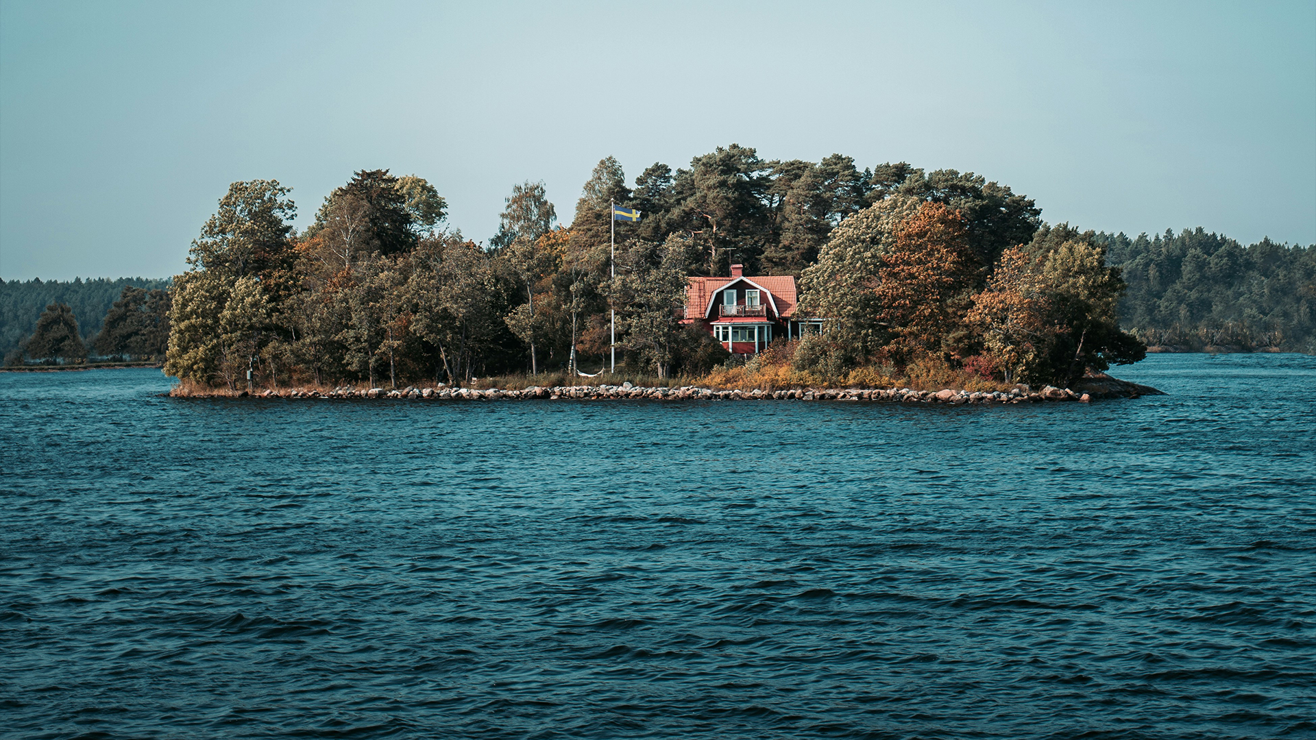 Bild på en liten ö med hus och flaggstång med svenska flaggan, i skärgården.
