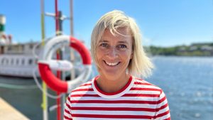 Leende, blond kvinna på en brygga. I bakgrunden en livboj.