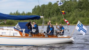 En äldre segelbåt går för motor. I aktersittbrunnen vinkar ett fler tal besättningsmedlemmar.