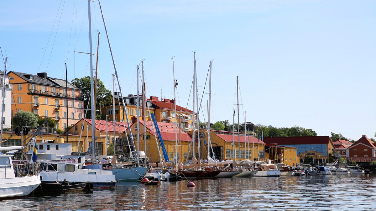 Varvsmiljö med småbåtar längs bryggor framför bostadshus.