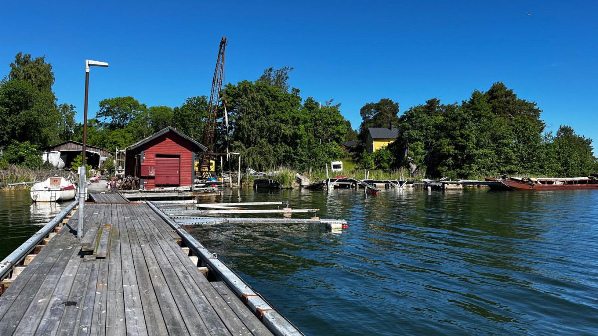 En nedgången brygga och en sjöbod. Man ser också en båthall, en lyftkran och en äldre segelbåt som ligger vid bryggan.