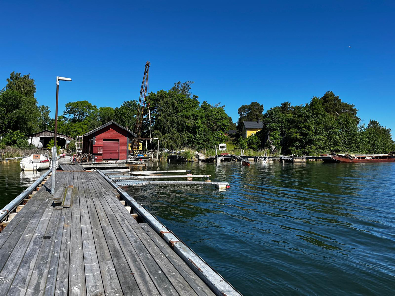 En nedgången brygga och en sjöbod. Man ser också en båthall, en lyftkran och en äldre segelbåt som ligger vid bryggan.