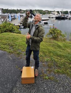 En man står framför en småbåtshamn. Han håller i och tittar i en sextant.