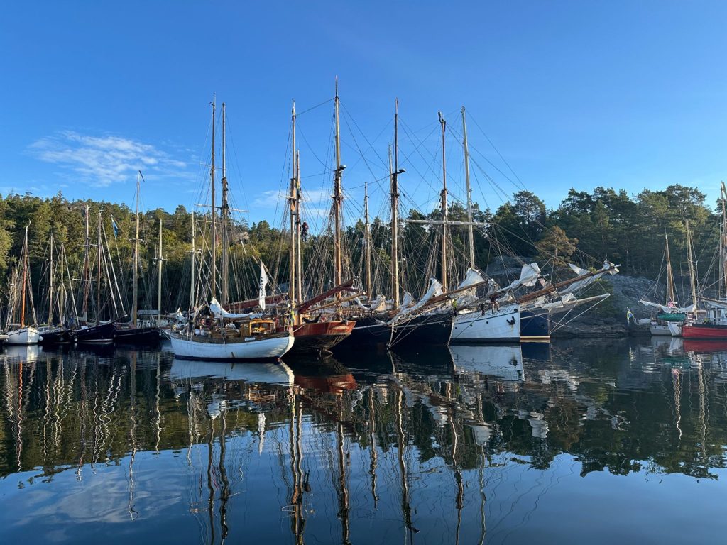 Ett antal segelskutor förtöjda i hamn. Vattnet ligger spegelblankt och himlen är blå.