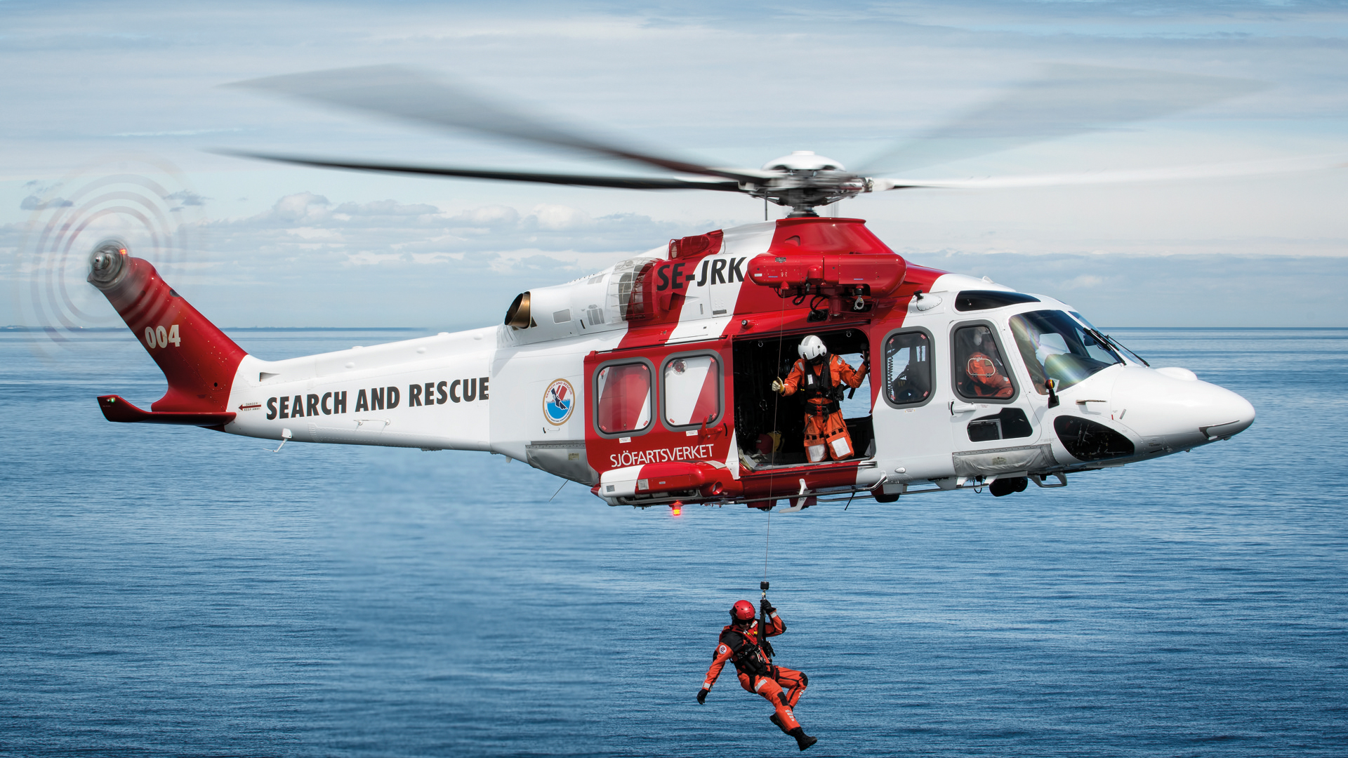 En räddningshelikopter hovrar över havet. Under helikoptern hänger en flygräddare.