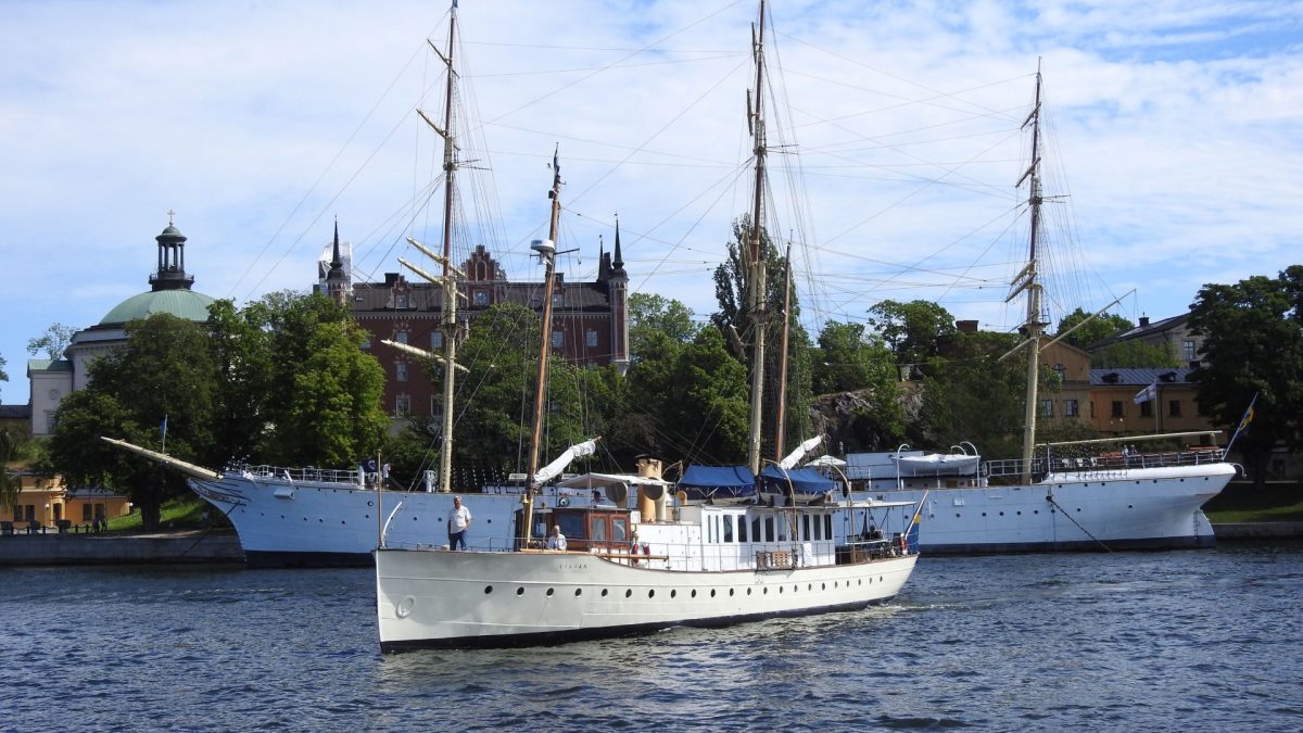 En äldre, lång träbåt med vitt skrov och vitmålad överbyggnad, framför skutan Af Chapman i Stockholm.